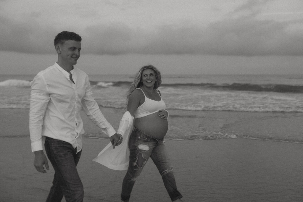 husband and wife walking on the beach looking at each other for their New Jersey Maternity photos