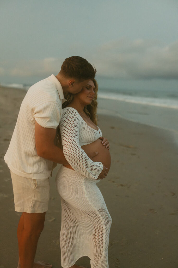 husband kissing wifes cheek and holding her belly for maternity photos in NJ