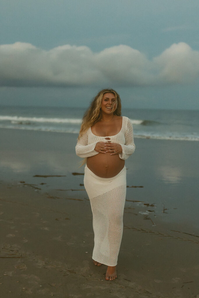 pregnant mama with her hands on her belly smiling at the camera on the beach in NJ